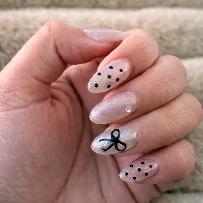 Uñas almendradas en blanco perlado con puntos negros pintados a mano, un lazo negro en el dedo medio y un cristal