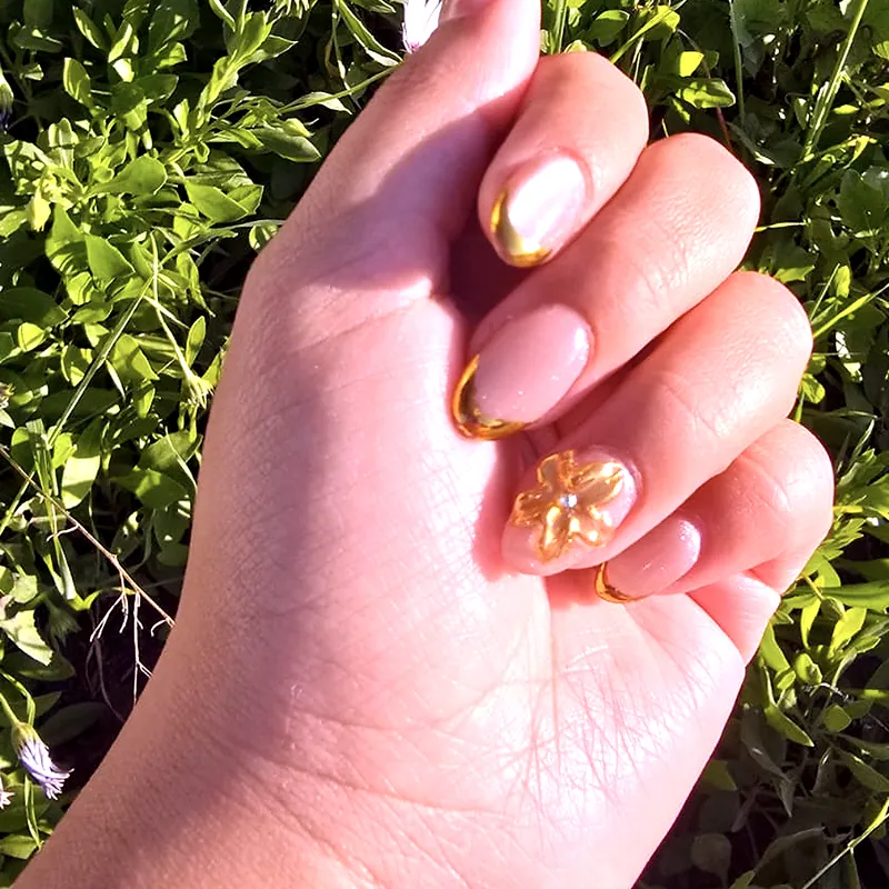 Uñas almendradas nude con French de línea dorada y una flor dorada en tres dimensiones sobre el anular, con un seto verde al fondo