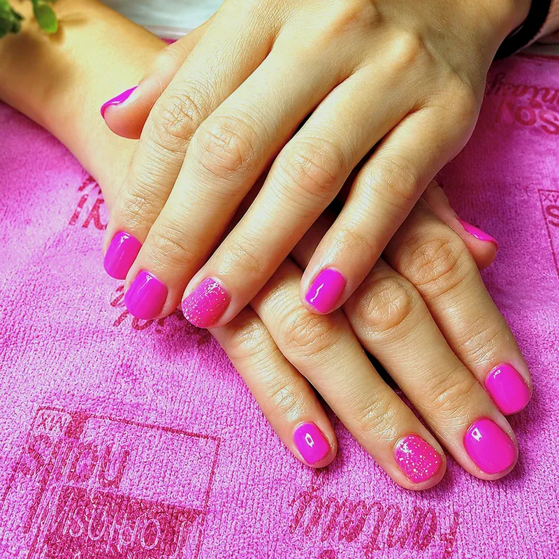 Dos manos con unas cortas en gel rosa fucsia y una uña con glitter, sobre una toalla rosa