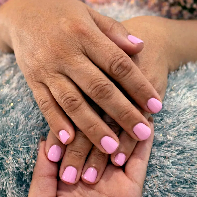 Dos manos con unas cortas y redondeadas en gel rosa pastel, apoyadas sobre un fondo plateado con flores