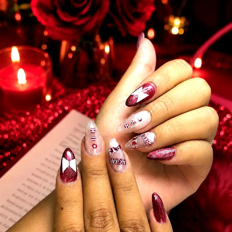 Uñas almendradas rojas y transparentes con glitter y letras pintadas a mano, entre velas encendidas y rosas rojas