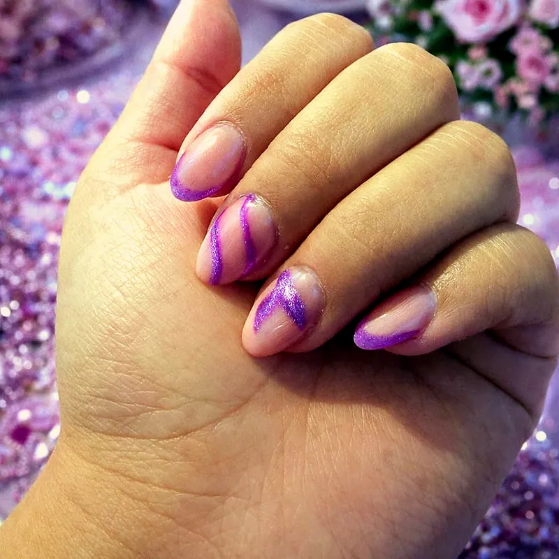 Uñas almendradas transparentes con remolinos violeta pintados a mano y glitter, sobre un fondo lila con rosas