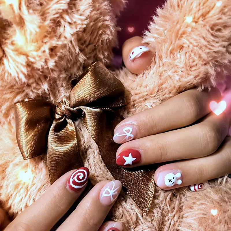 Uñas cortas almendradas con dibujos de invierno pintados a mano —un remolino de caramelo, una estrella, una carita de osito y lacitos— sobre piel sintética beige